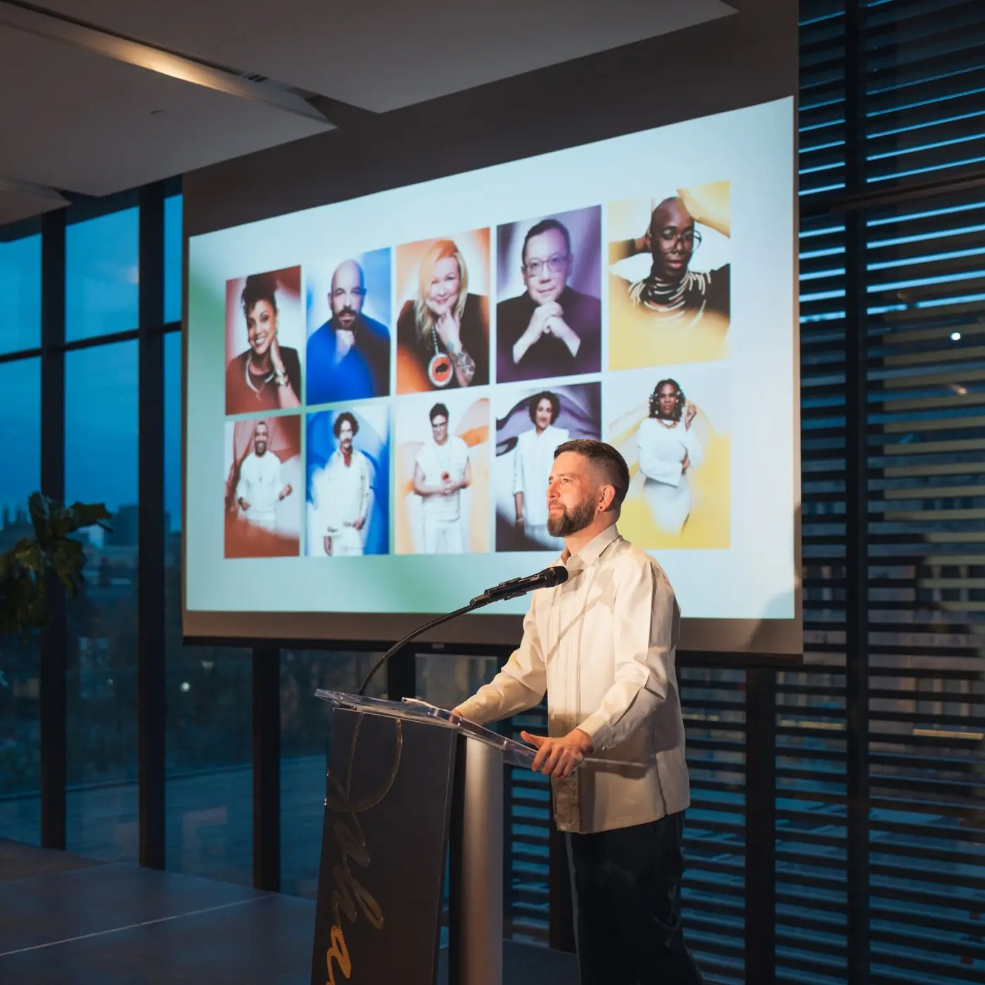 A speaker at a podium during The Johannas awards, with winner portraits projected behind